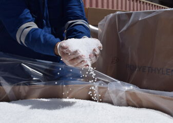 Hände in Arbeitshandschuhen halten und streuen weiße Kunststoffpellets über eine mit Folie ausgelegte Kiste mit der Aufschrift 'POLYETHYLENE'