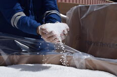 Hände in Arbeitshandschuhen halten und streuen weiße Kunststoffpellets über eine mit Folie ausgelegte Kiste mit der Aufschrift 'POLYETHYLENE'